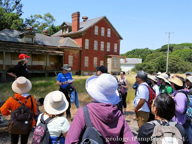 A group of people looking as a man talks, an old brick building in the background